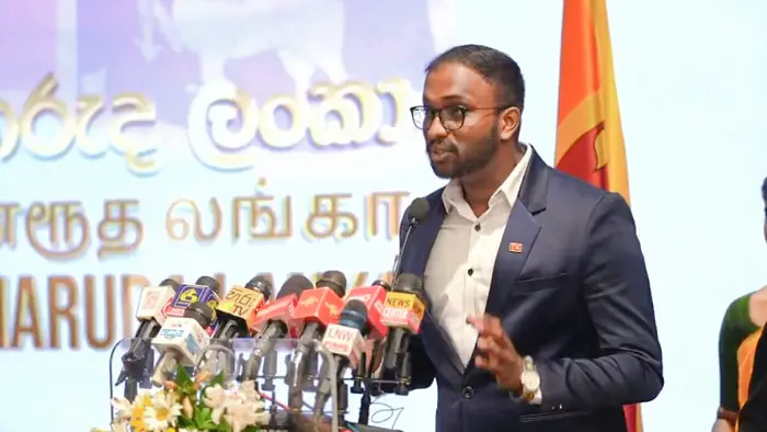 Mayantha speaking at a podium in front of press microphones